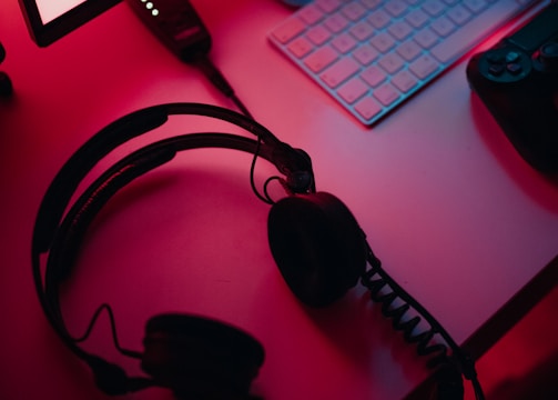 A dimly lit gaming setup with a monitor displaying a game's menu on the screen. A mechanical keyboard sits in front of the monitor. A pair of over-ear headphones and a game controller rest on the desk. The scene is bathed in a red and blue light, creating a moody gaming atmosphere.