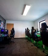 A well-lit gaming room features multiple gaming stations with high-backed chairs and monitors. The room has a minimalist setup with green and black color tones. Large posters adorn the walls, and the room appears clean and organized.