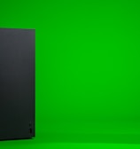 A black, rectangular gaming console stands upright against a vibrant green background. The console has a simple, sleek design with minimalistic features including a disc slot and a few buttons.