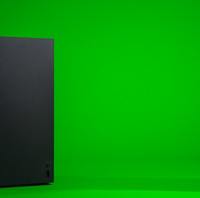 A black, rectangular gaming console stands upright against a vibrant green background. The console has a simple, sleek design with minimalistic features including a disc slot and a few buttons.