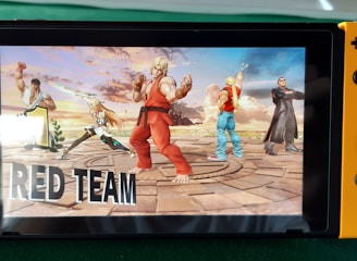 A handheld gaming console displays a screenshot of a fighting game featuring cartoonish characters in dynamic poses. The characters are standing in a virtual arena setting with a sky and cloud backdrop. The console is placed on a green surface.