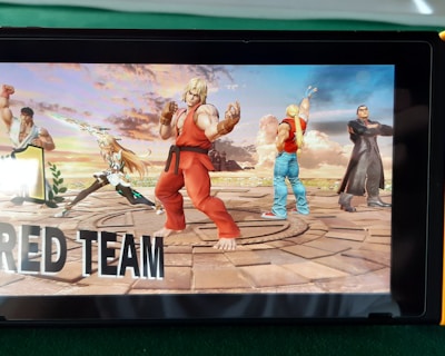 A handheld gaming console displays a screenshot of a fighting game featuring cartoonish characters in dynamic poses. The characters are standing in a virtual arena setting with a sky and cloud backdrop. The console is placed on a green surface.