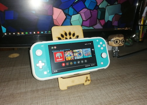 A turquoise handheld gaming console is placed on a wooden stand in front of a computer monitor displaying geometric shapes. To the right, there is a small figurine of a person wearing glasses and a suit. The gaming console screen shows a menu with various game icons.