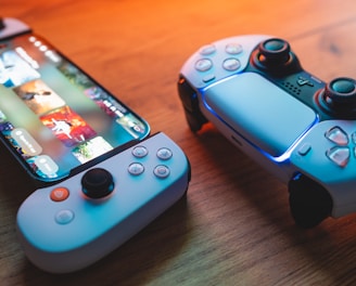 A gaming scenario featuring a smartphone with a game interface next to two controllers on a wooden surface. The smartphone displays vibrant images and icons, while the controllers feature a sleek, modern design with buttons and joysticks prominently visible. Warm lighting enhances the scene.