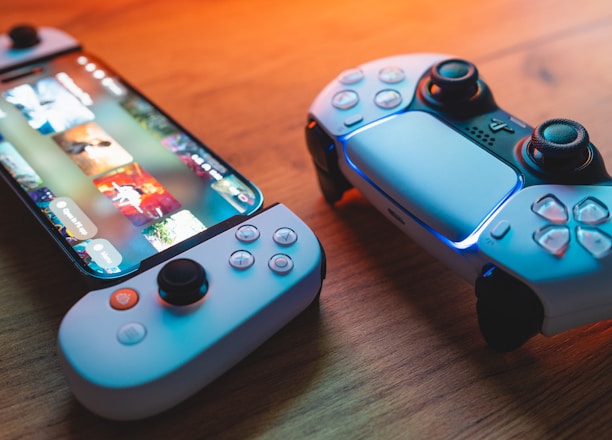 A gaming scenario featuring a smartphone with a game interface next to two controllers on a wooden surface. The smartphone displays vibrant images and icons, while the controllers feature a sleek, modern design with buttons and joysticks prominently visible. Warm lighting enhances the scene.