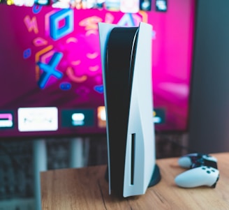 A gaming console is placed on a wooden table with a game controller nearby. In the background, a television screen displays colorful graphics, including geometric shapes and vibrant colors.