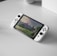 A portable gaming console with a screen showing a video game landscape, resting on a light-colored surface. The console has white controllers attached to either side of the screen. Nearby, there seems to be a white docking station or accessory.