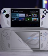 A handheld gaming console with a screen displaying a game library menu is placed on top of its packaging. The console features various buttons, a joystick, and a circular light around the left joystick. The background is blurred with purple, yellow, and blue tones.