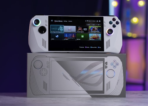 A handheld gaming console with a screen displaying a game library menu is placed on top of its packaging. The console features various buttons, a joystick, and a circular light around the left joystick. The background is blurred with purple, yellow, and blue tones.