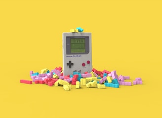 A vintage handheld gaming console surrounded by colorful, block-like pieces reminiscent of a classic puzzle game. The console has a small screen displaying a game in progress, with two red buttons and a directional pad. The vibrant pieces are scattered around it, creating a playful and nostalgic atmosphere.