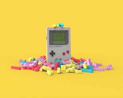 A vintage handheld gaming console surrounded by colorful, block-like pieces reminiscent of a classic puzzle game. The console has a small screen displaying a game in progress, with two red buttons and a directional pad. The vibrant pieces are scattered around it, creating a playful and nostalgic atmosphere.