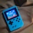 A handheld gaming device with a bright blue casing rests on a soft, dark fabric background. The screen displays a pixelated video game scene with options to select either Chinese or English language. The layout includes a classic D-pad and buttons, reminiscent of retro gaming consoles.