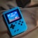 A handheld gaming device with a bright blue casing rests on a soft, dark fabric background. The screen displays a pixelated video game scene with options to select either Chinese or English language. The layout includes a classic D-pad and buttons, reminiscent of retro gaming consoles.